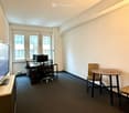 Minimalistic office room with desks, monitors, and chairs. A television on a stand and a small table with another chair. White walls and black carpet. A window with a city view.