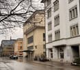 A building with a balcony and arched windows, next to a building with large windows, on a rainy street with cars and pedestrians.