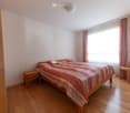 Bedroom with a double bed, wooden nightstand, wooden chair, white walls, wooden floor, lamp, large window with curtains