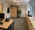 Furnished Room. An office space with a desk, computer monitor, telephone, and keyboard. A cabinet and plants are on the right side, and a coat is hanging on the wall.