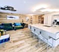 Open-plan living and kitchen area with wooden flooring, wooden wall, modern furniture, large island with bar stools, kitchen appliances, and a dining table.