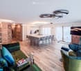Open-plan living and dining area with wooden floor, wooden wall, large windows, dining table with chairs, kitchen, TV, sofa, armchairs, shelves, magazines on the table