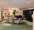 Empty reception area with reception desks, plants, monitors, and carpeted floor.