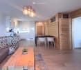Open living room with dining area, wooden floor, wooden walls, modern furniture, kitchen with appliances, couch, coffee table, rug, and decorative items