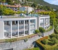 modern residential building with multiple balconies, glass facade, roof terrace, set against a scenic hillside background