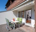 Outdoor dining area with table and chairs, flower vase, wooden floor, white wall, wooden railing, open door, wooden balcony