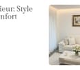 Modern living room with white couch, coffee table, and white rug. Vase with flowers on the table, books, and pillows on the couch. Picture frame on the wall.