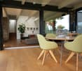 Dining area with wooden floor, large windows, round table, green chairs, flower vase, couch, staircase, plants