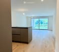 Empty room with light wooden flooring, white walls, and a small kitchen counter in the corner. Large sliding glass doors lead to an outdoor area.