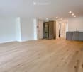 Empty room with light wooden flooring, white walls, and a small kitchen area visible in the background.