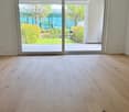 Empty room with light wooden flooring and large sliding glass doors leading to a patio area with greenery visible outside.