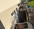 Raised ground floor, beige wall, red shutters, small window, concrete stairs, wooden bench, wooden fence, plants, grass
