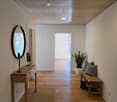 Empty hallway with wooden floor, white walls, wooden bench, round mirror, potted plant, rug, and ceiling lights
