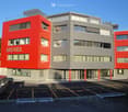 A modern multi-story motel building with a red and gray exterior, large windows, and a parking lot in front.
