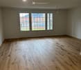 A spacious empty room with wooden floors, glass sliding doors, three windows, a white ceiling, and a ceiling light.