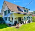 2 story house, white exterior, brown roof, green shutters, large front yard, patio with seating, garden, awning