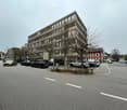 Office building with multiple windows, cars parked in front, street lights, trees, and a person walking