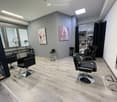A salon with black leather chairs, a wooden floor, a gray wall, a shelf with bottles, and two paintings on the wall.