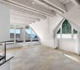 Bright, airy attic space with exposed wooden beams, large windows, and a fireplace. The room features a tiled floor and has a view of the sea through the windows.