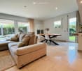 modern living room with large windows, beige sofa, wooden table, white chairs, beige carpet, white tiles, white cabinets, plants