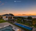 A modern house with a spacious outdoor area featuring a swimming pool, hot tub, wooden deck, and well-maintained lawn, overlooking a scenic lake and mountain view at sunset.