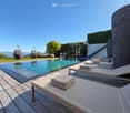 A modern outdoor area with a rectangular swimming pool, wooden decking, lounge chairs, and towels, surrounded by greenery and a clear blue sky.