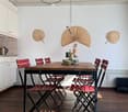Dining room with wooden table, red chairs, white walls, wooden floor, hanging lamps, kitchen cabinets, decorative fans on the wall