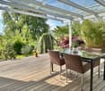 Wooden deck with a glass table and chairs, covered by a glass pergola, surrounded by plants and trees, with a view of the countryside