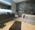 Modern bathroom with marble walls, wooden floor, large mirror, white sink, and bathtub with towels.