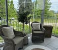 Balcony / Terrace. A balcony with a view of trees and a road, featuring two wicker chairs with cushions, a potted plant, a storage box, and a round rug.