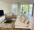 Living Room. Modern living room with a large L-shaped sofa, a wooden coffee table, and a flat-screen TV on an entertainment center. A decorative pillow and a throw pillow are on the sofa. A rug covers part of the wooden floor. The room has large glass doo