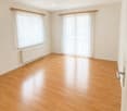 Empty room with wooden floors, white walls, and two large windows with white curtains. A white radiator is mounted on the wall below one of the windows.