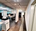 Modern office space with a desk, chairs, multiple screens, a printer, and a small kitchen area visible in the background.