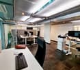Modern office space with desks, chairs, computers, and a printer. Features a sleek design with glass partitions and a plant in the corner.