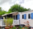 A small, single-story mobile home with light beige siding and blue shutters, situated in a lush, green garden. The home features a covered porch area with a white railing, potted plants, and a small decorative statue. The surrounding area includes trees a