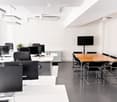 Modern office space with multiple workstations, a conference table, and a large screen. The room features sleek black chairs, desks, and a potted plant, with a polished dark floor and white walls.