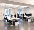 Modern office space with white desks, black office chairs, multiple computer setups, and a coat rack near the entrance.
