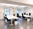 Modern office space with white tables, black chairs, trash bins, and a coat rack near the window.