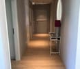 A modern hallway with light wooden flooring, white walls, and several doors leading to different rooms. A small table with a lamp is placed against the right wall.