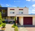 House. Modern single-story house with light beige walls, dark red garage door, and small garden area with potted plants and gravel pathway.