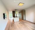 Modern kitchen with light wood flooring, white island, stainless steel range hood, and built-in cabinets.