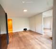 Empty room with light wooden flooring, white walls, and a single black stool near the center. A doorway leads to another brightly lit room.
