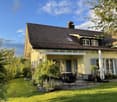 House. Two-story yellow house with a dark roof, multiple windows, and a covered porch. The garden features a patio with outdoor furniture, including a table and chairs, surrounded by greenery and a well-maintained lawn.