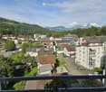 Overlook of town with many buildings, cars parked on roads, hills covered in trees, mountains in the distance.