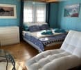 Bedroom. Cozy bedroom with wooden walls, a large bed with pillows and a blue comforter, a white nightstand, a white armchair, a glass table, and two picture frames on the walls.