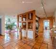 Corridor / Hallway. Spacious hallway with terracotta tiles, wooden shelving unit, decorative statue, and a small table with a bowl and flowers. The hallway leads to a living area with a potted plant and seating area.