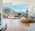 Furnished Room. Bright living room with large windows offering a scenic mountain view, featuring a white sofa, a round table with chairs, a floor lamp, a television, and a bookshelf.