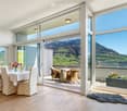Furnished Room. Bright dining area with large windows offering scenic mountain views, featuring a dining table with chairs, a flower vase, and a sideboard with table lamps.