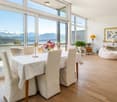 Furnished Room. Bright dining area with large windows offering a scenic mountain view, featuring a dining table with chairs, table lamps, potted plants, a sofa, and a picture frame on the wall.