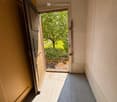 Corridor / Hallway. A narrow hallway with a grey carpeted floor, beige walls, and an open door leading to a garden area with trees and greenery.